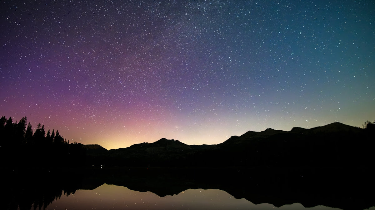 Starry night sky with colorful glow over mountains and lake.