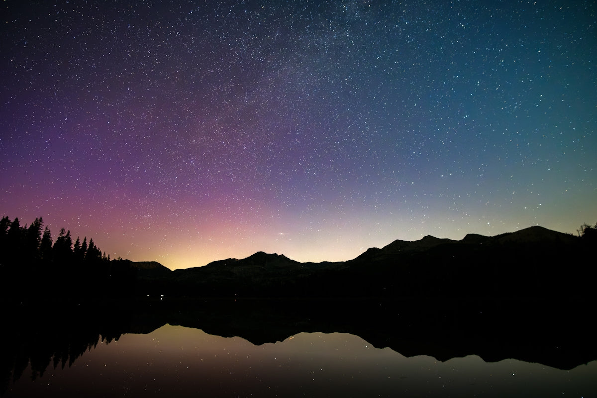 Starry night sky with colorful glow over mountains and lake.